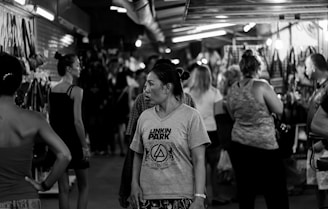 People are walking and browsing in a crowded indoor market. The environment is dimly lit, with various stalls lining the area, displaying items like bags and clothes. Some individuals are wearing casual attire, and one person is wearing a distinctive band t-shirt.