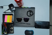 A collection of various office and personal items is arranged neatly on a light surface. There is a spiral-bound notebook with a pair of black sunglasses placed on it. Earphones are also on top of the notebook. Next to it, a smartphone is plugged in and charging. A compact wireless mouse is positioned nearby. A storage box holds several stationery items including pens, highlighters, and sticky notes in pink, orange, and yellow colors. A set of keys and a wireless internet dongle are also part of the arrangement.