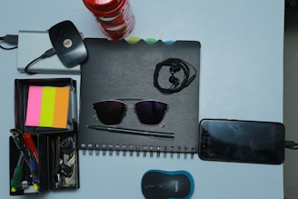 A collection of various office and personal items is arranged neatly on a light surface. There is a spiral-bound notebook with a pair of black sunglasses placed on it. Earphones are also on top of the notebook. Next to it, a smartphone is plugged in and charging. A compact wireless mouse is positioned nearby. A storage box holds several stationery items including pens, highlighters, and sticky notes in pink, orange, and yellow colors. A set of keys and a wireless internet dongle are also part of the arrangement.