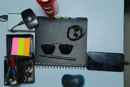 A collection of various office and personal items is arranged neatly on a light surface. There is a spiral-bound notebook with a pair of black sunglasses placed on it. Earphones are also on top of the notebook. Next to it, a smartphone is plugged in and charging. A compact wireless mouse is positioned nearby. A storage box holds several stationery items including pens, highlighters, and sticky notes in pink, orange, and yellow colors. A set of keys and a wireless internet dongle are also part of the arrangement.