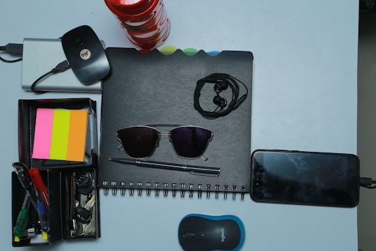 A collection of various office and personal items is arranged neatly on a light surface. There is a spiral-bound notebook with a pair of black sunglasses placed on it. Earphones are also on top of the notebook. Next to it, a smartphone is plugged in and charging. A compact wireless mouse is positioned nearby. A storage box holds several stationery items including pens, highlighters, and sticky notes in pink, orange, and yellow colors. A set of keys and a wireless internet dongle are also part of the arrangement.