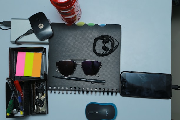 A collection of various office and personal items is arranged neatly on a light surface. There is a spiral-bound notebook with a pair of black sunglasses placed on it. Earphones are also on top of the notebook. Next to it, a smartphone is plugged in and charging. A compact wireless mouse is positioned nearby. A storage box holds several stationery items including pens, highlighters, and sticky notes in pink, orange, and yellow colors. A set of keys and a wireless internet dongle are also part of the arrangement.