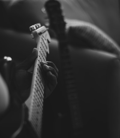 A hand gripping the neck of an electric guitar, focusing on the strings and frets. The background is blurred, giving a sense of depth. The lighting is soft, casting gentle shadows.