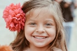 A young girl with light brown hair smiles broadly, displaying a joyful expression. A large, vibrant pink flower adorns her hair on the left side, adding a playful touch. The background is softly blurred, emphasizing her cheerful face.