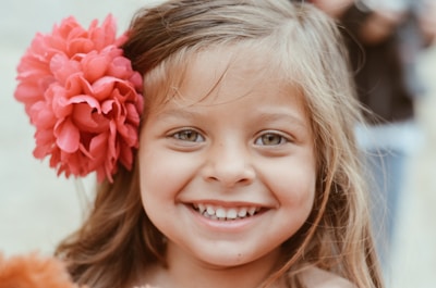 A joyful little girl wearing a bright pink bow smiling outdoors in natural light.