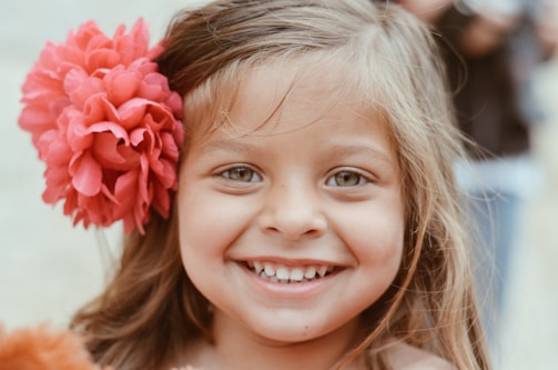A young girl with light brown hair smiles broadly, displaying a joyful expression. A large, vibrant pink flower adorns her hair on the left side, adding a playful touch. The background is softly blurred, emphasizing her cheerful face.