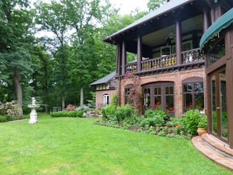 An elegant two-story brick house is surrounded by lush green grass and vibrant gardens. The house features arched windows and a covered balcony with wooden railings. Climbing plants and flowers provide a colorful accent against the brick walls. In the garden, there is a decorative stone sculpture or birdbath standing on a pedestal, adding to the picturesque scene. Tall trees frame the background, creating a serene and secluded atmosphere.