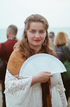 A young woman with long wavy hair, wearing earrings and bright red lipstick, is holding a white handheld fan. She is dressed in a white lace garment with a brown shawl. The background includes several blurred figures and a body of water.