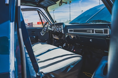 The interior of an old truck with a classic dashboard design is visible, featuring a steering wheel and well-worn seats. Sunlight casts shadows across the seats, highlighting the aging fabric and the details of the dashboard. Outside, parts of a car dealership with signage are visible through the vehicle's open door.