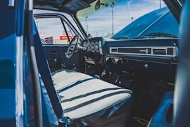 The interior of an old truck with a classic dashboard design is visible, featuring a steering wheel and well-worn seats. Sunlight casts shadows across the seats, highlighting the aging fabric and the details of the dashboard. Outside, parts of a car dealership with signage are visible through the vehicle's open door.