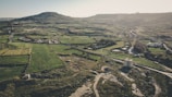 An aerial view of a vast rural landscape featuring patchwork fields and agricultural land, interspersed with a few buildings and roads. A prominent hill rises in the background, casting shadows over the terrain. The scene evokes a serene, pastoral atmosphere with well-defined field boundaries and a mix of green and earthy tones.