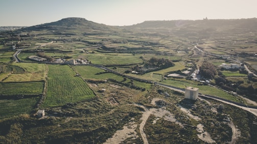 An aerial view of a vast rural landscape featuring patchwork fields and agricultural land, interspersed with a few buildings and roads. A prominent hill rises in the background, casting shadows over the terrain. The scene evokes a serene, pastoral atmosphere with well-defined field boundaries and a mix of green and earthy tones.