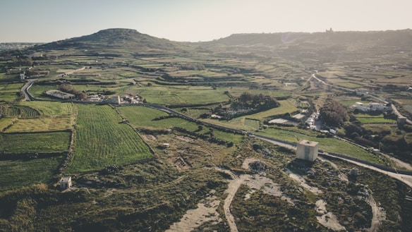 An aerial view of a vast rural landscape featuring patchwork fields and agricultural land, interspersed with a few buildings and roads. A prominent hill rises in the background, casting shadows over the terrain. The scene evokes a serene, pastoral atmosphere with well-defined field boundaries and a mix of green and earthy tones.
