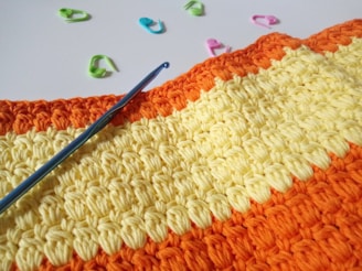 Colorful crochet work with alternating stripes of orange and yellow yarn. A blue crochet hook is placed on the work. Surrounding the crochet piece, there are several plastic stitch markers in pink, blue, and green colors.
