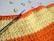 Colorful crochet work with alternating stripes of orange and yellow yarn. A blue crochet hook is placed on the work. Surrounding the crochet piece, there are several plastic stitch markers in pink, blue, and green colors.