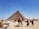 A large pyramid stands majestically under a clear blue sky. In the foreground, several people interact around the base of the pyramid, with some riding camels adorned in colorful blankets. To the left, a market stall displays various souvenirs and trinkets. The ground is sandy and the atmosphere suggests a warm, sunny day.