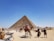 A large pyramid stands majestically under a clear blue sky. In the foreground, several people interact around the base of the pyramid, with some riding camels adorned in colorful blankets. To the left, a market stall displays various souvenirs and trinkets. The ground is sandy and the atmosphere suggests a warm, sunny day.