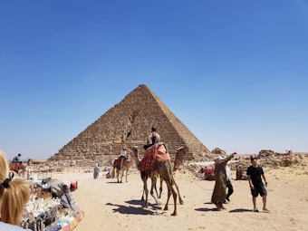 A large pyramid stands majestically under a clear blue sky. In the foreground, several people interact around the base of the pyramid, with some riding camels adorned in colorful blankets. To the left, a market stall displays various souvenirs and trinkets. The ground is sandy and the atmosphere suggests a warm, sunny day.