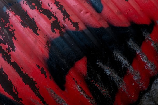 A section of corrugated metal covered in red and black spray paint, with the paint pattern resembling abstract shapes or perhaps an animal silhouette. The red paint is applied unevenly, allowing the dark color of the metal to show through in places, creating a distressed and textured appearance.