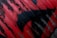 A section of corrugated metal covered in red and black spray paint, with the paint pattern resembling abstract shapes or perhaps an animal silhouette. The red paint is applied unevenly, allowing the dark color of the metal to show through in places, creating a distressed and textured appearance.