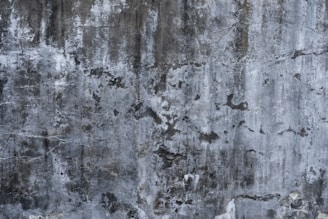 A textured concrete wall with a weathered and aged appearance, showing various shades of gray and black with subtle hints of white. The surface is marked by rough patches, cracks, and stains, giving it a rugged, industrial feel.