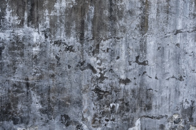 A textured concrete wall with a weathered and aged appearance, showing various shades of gray and black with subtle hints of white. The surface is marked by rough patches, cracks, and stains, giving it a rugged, industrial feel.