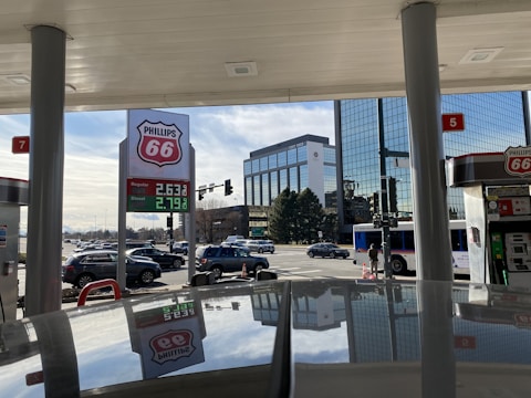 A Phillips 66 gas station with a view of the price sign showing regular and diesel prices. Several cars are seen on the street, and a public bus is at a traffic light. The background features modern glass office buildings and a clear sky.