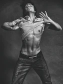 A black and white photograph of a shirtless man with a muscular build, posed against a textured backdrop. The man is depicted in motion, lifting his shirt above his chest. He wears jeans and his hair is slightly tousled, adding to the dynamic and expressive quality of the image.