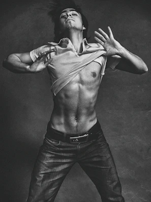 A black and white photograph of a shirtless man with a muscular build, posed against a textured backdrop. The man is depicted in motion, lifting his shirt above his chest. He wears jeans and his hair is slightly tousled, adding to the dynamic and expressive quality of the image.