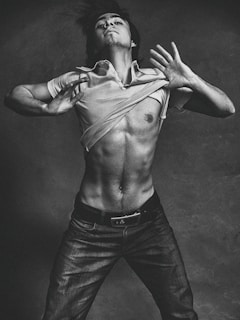 A black and white photograph of a shirtless man with a muscular build, posed against a textured backdrop. The man is depicted in motion, lifting his shirt above his chest. He wears jeans and his hair is slightly tousled, adding to the dynamic and expressive quality of the image.