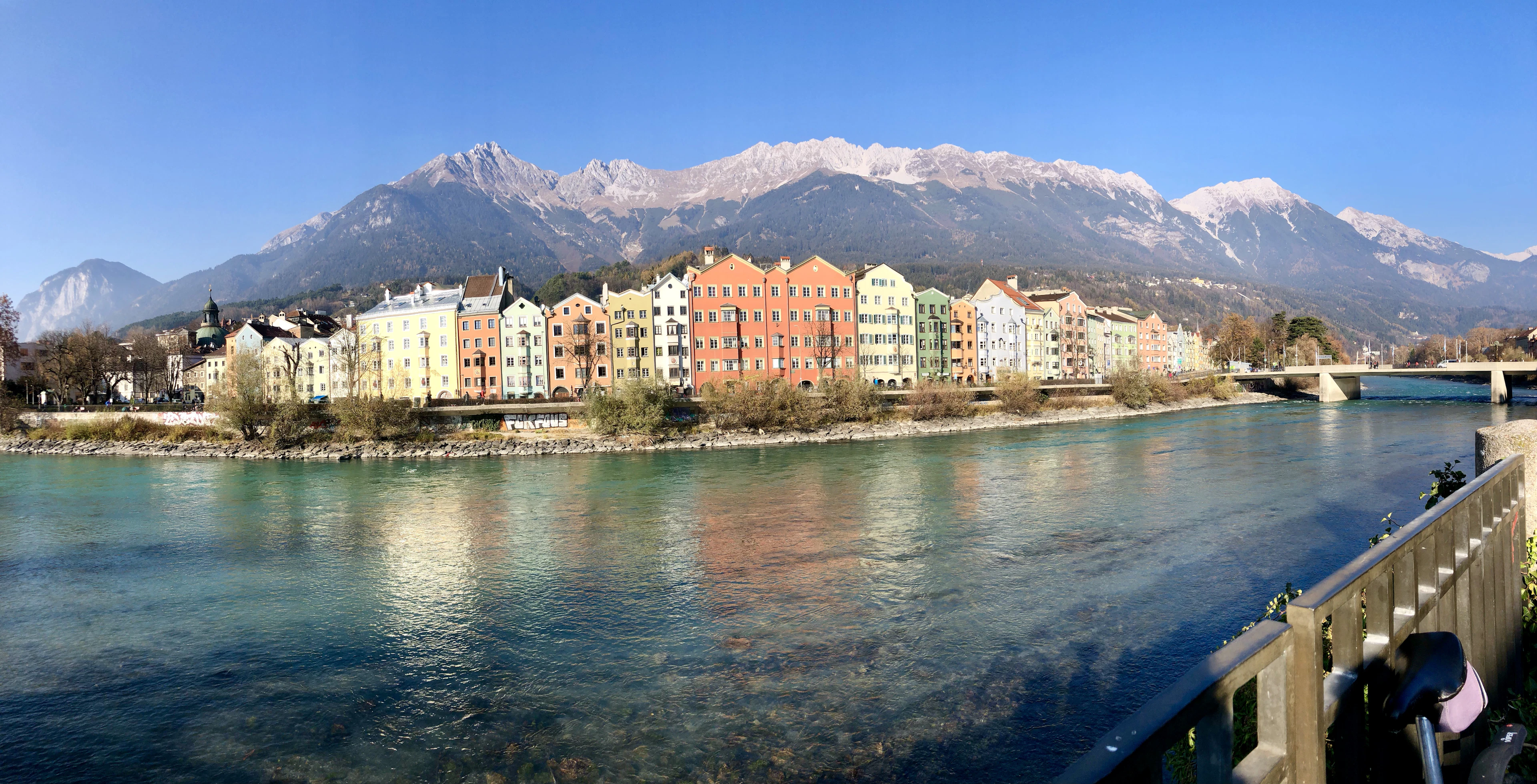 Colorful Mariahilf houses lining the Inn River in Innsbruck with a bridge and snow-covered mountains behind.