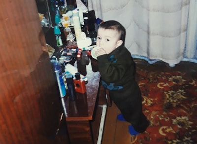 A small child stands near a cluttered dressing table filled with various cosmetic items and toiletries, including bottles, brushes, and makeup palettes. The child is wearing a dark sweater and pants, and is gazing curiously at the camera. The room has a floral-patterned carpet and light-colored drapes.