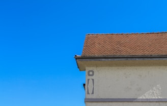 A section of a building with a beige stucco wall and a steeply pitched roof covered in brown shingles. The sky is a clear, vibrant blue. The shadow of a person or object is visible on the wall in the lower left.