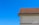 A section of a building with a beige stucco wall and a steeply pitched roof covered in brown shingles. The sky is a clear, vibrant blue. The shadow of a person or object is visible on the wall in the lower left.