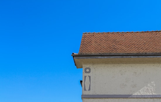 A section of a building with a beige stucco wall and a steeply pitched roof covered in brown shingles. The sky is a clear, vibrant blue. The shadow of a person or object is visible on the wall in the lower left.