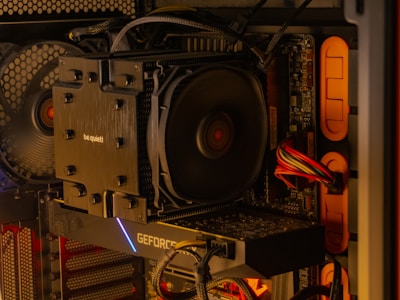 A close-up of an open computer case reveals internal components, including a large CPU cooler labeled 'be quiet!', a graphics card with the label 'GEFORCE', and neatly arranged cables. The lighting inside the case emits a warm, orange glow, highlighting the fan and circuit board details.