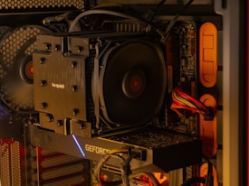 A close-up of an open computer case reveals internal components, including a large CPU cooler labeled 'be quiet!', a graphics card with the label 'GEFORCE', and neatly arranged cables. The lighting inside the case emits a warm, orange glow, highlighting the fan and circuit board details.