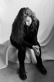 A person with long hair is sitting with a bowed head, clasping their hands together. The image is in black and white, creating a somber and reflective atmosphere. The backdrop is a simple draped fabric, and the person is dressed in dark clothing with large boots.
