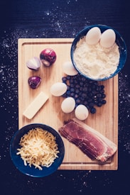 A wooden chopping board is surrounded by a dark, starry background. On the board, there are ingredients including eggs, flour in a bowl, whole red onions, a block of cheese, shredded cheese in a separate bowl, a stack of bacon slices, and a cluster of dark berries.
