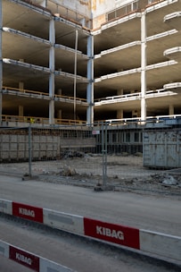An incomplete multi-story concrete structure is visible, with exposed floors and support columns. There is construction fencing in front, along with red and white barricades labeled KIBAG. The area is dusty with construction debris and materials scattered around.