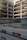 An incomplete multi-story concrete structure is visible, with exposed floors and support columns. There is construction fencing in front, along with red and white barricades labeled KIBAG. The area is dusty with construction debris and materials scattered around.
