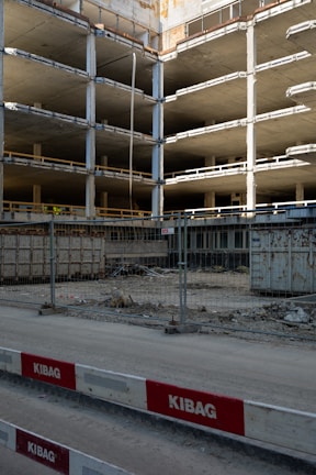 An incomplete multi-story concrete structure is visible, with exposed floors and support columns. There is construction fencing in front, along with red and white barricades labeled KIBAG. The area is dusty with construction debris and materials scattered around.