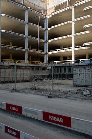 An incomplete multi-story concrete structure is visible, with exposed floors and support columns. There is construction fencing in front, along with red and white barricades labeled KIBAG. The area is dusty with construction debris and materials scattered around.
