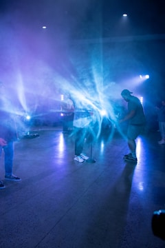 A group of people is gathered on a stage with a misty atmosphere, illuminated by blue and purple stage lights. The individuals are engaged in a musical performance or rehearsal, standing around microphones and instruments.