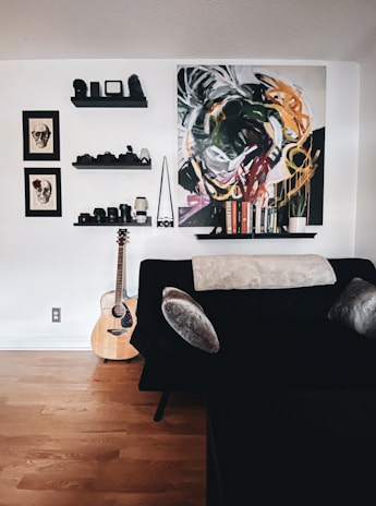 A cozy room featuring a black couch with grey cushions, accompanied by a beige throw. On the wall, there are three floating shelves displaying cameras, lenses, and decor items. Two framed skull artworks hang beside a colorful abstract painting. A wooden acoustic guitar rests against the wall, placed on a hardwood floor.