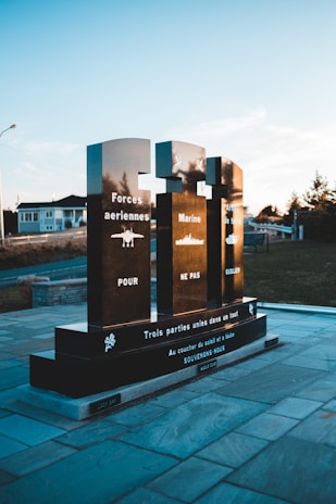 A memorial structure with three black granite pillars displaying white inscriptions honoring the different branches of the military. The words 'Forces aériennes,' 'Marine,' and 'Armée de terre' are inscribed alongside icons representing an airplane, a ship, and a tank respectively. The monument is situated in an outdoor setting with a paved stone path and some trees and buildings in the background, with a clear blue sky above.