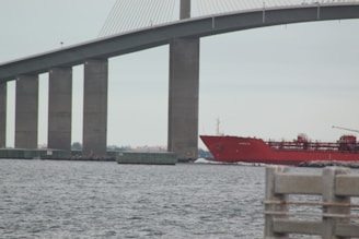 A large, red cargo ship passes under a massive bridge with tall vertical supports. The water appears calm, and there is a solid structure in the foreground, possibly part of a dock or pier.