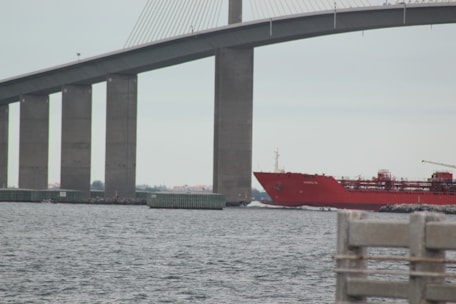 A large, red cargo ship passes under a massive bridge with tall vertical supports. The water appears calm, and there is a solid structure in the foreground, possibly part of a dock or pier.