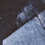 The image depicts a close-up view of a textured concrete surface with chalk markings or writings. There is a prominent crack running through the concrete, adding a sense of ruggedness. The lighting is dim, and the colors are predominantly cool tones, giving it a moody atmosphere.