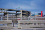 An urban landscape featuring multiple overpasses and highway bridges. The foreground shows a concrete barrier with metal railings, while towering over the scene are elevated roadways. In the background, there are city buildings, notably including a prominent red cylinder-shaped structure next to modern glass and steel buildings.